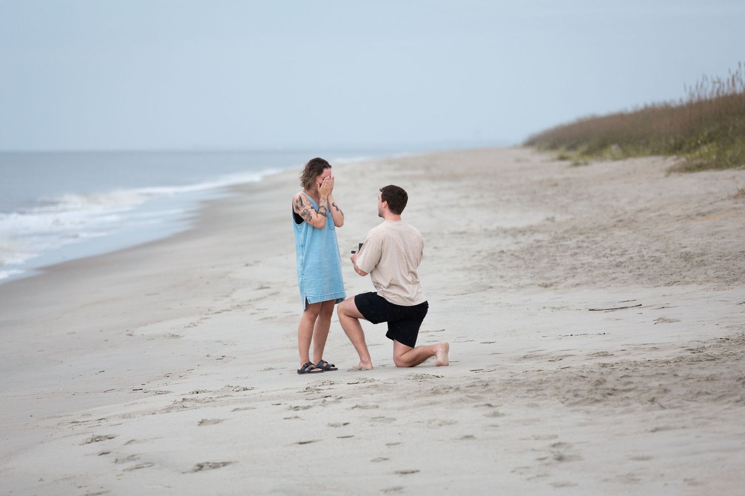 A romantic proposal moment unfolds on a serene beach with waves and sand dunes in the background.