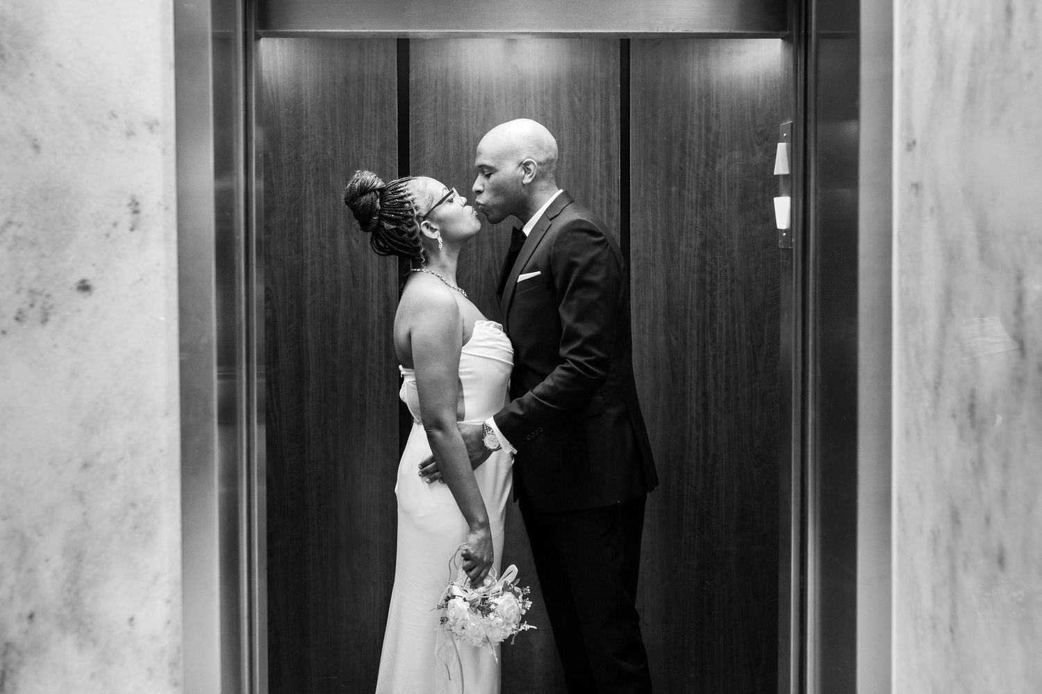 Black and white photo of a bride and groom sharing a kiss in an elevator on their wedding day.
