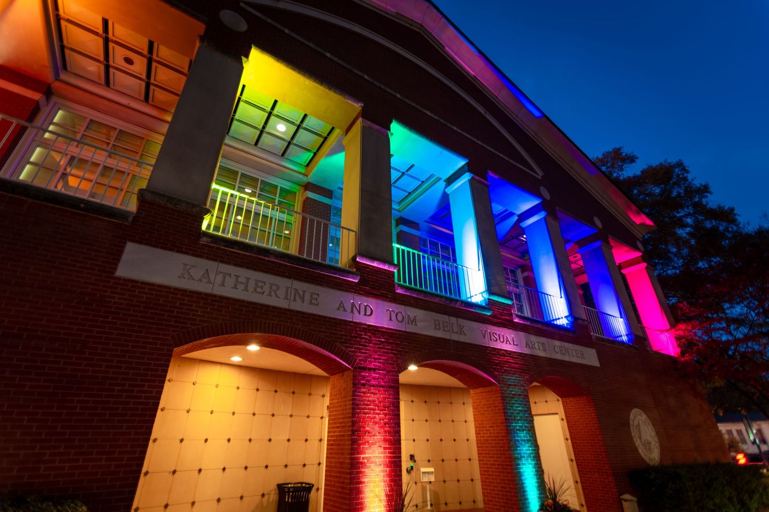 Building exterior illuminated with colorful rainbow LED lighting against a dark blue evening sky.