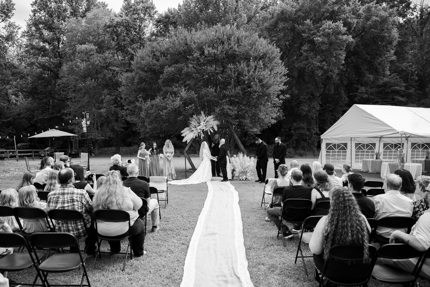 Wedding guests gather outdoors under trees at a casual garden reception with white tents and chairs.