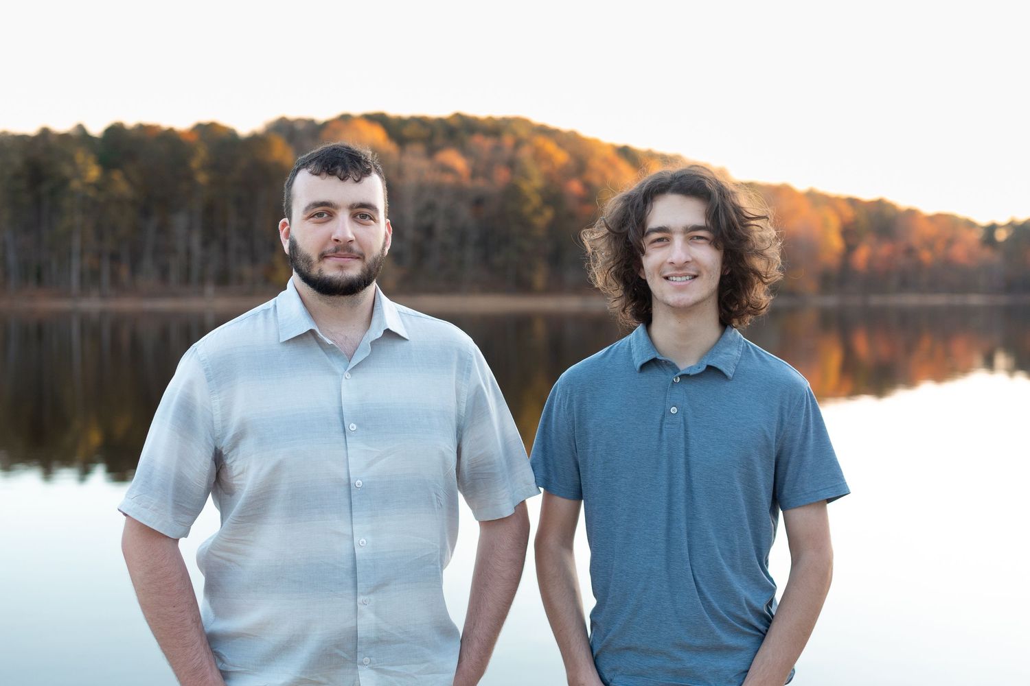 Two people stand together smiling against a scenic autumn lake backdrop at sunset.