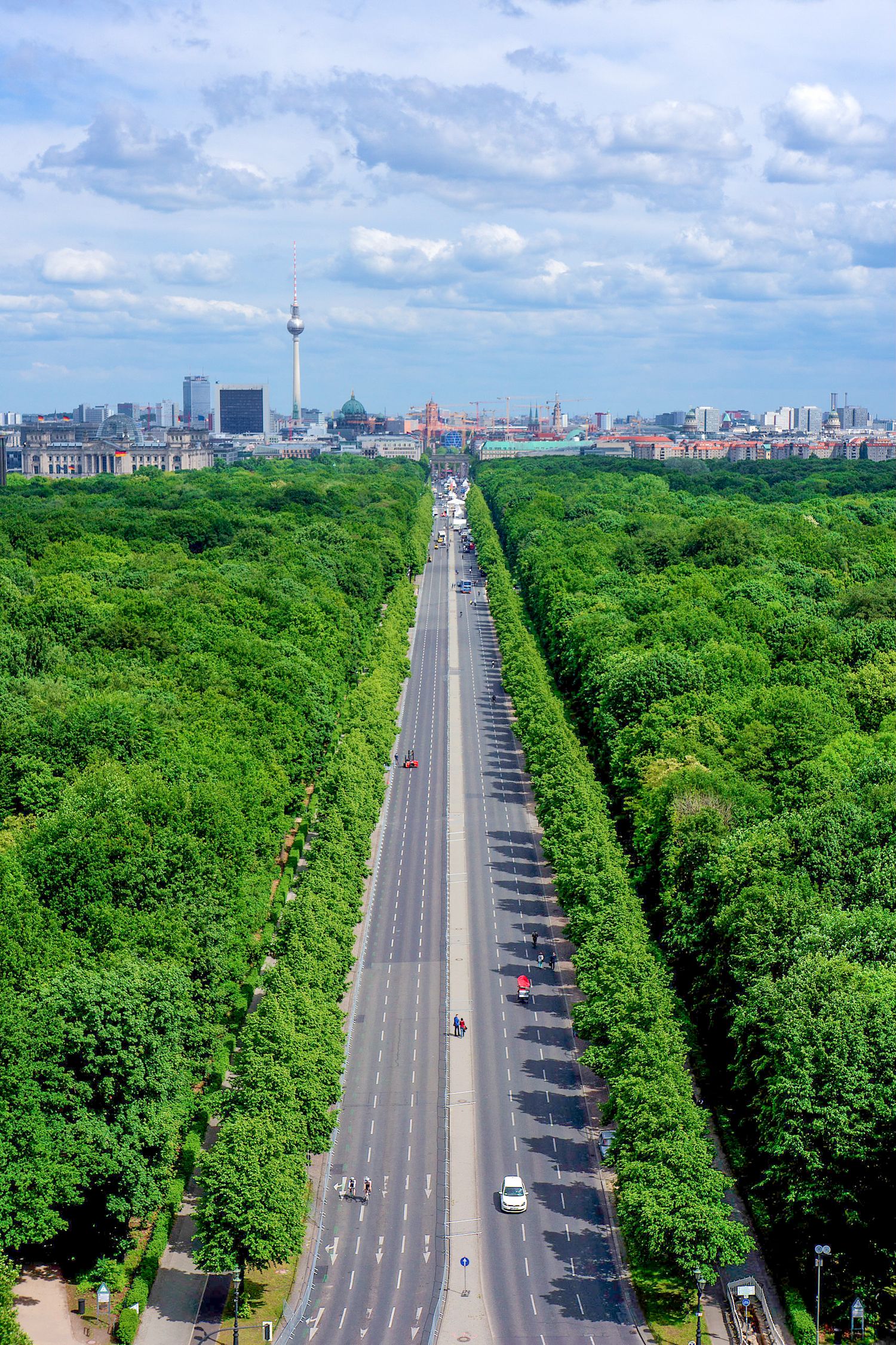 Aerial view of a long straight road flanked by lush green trees leading to the Berlin city skyline under a cloudy sky.