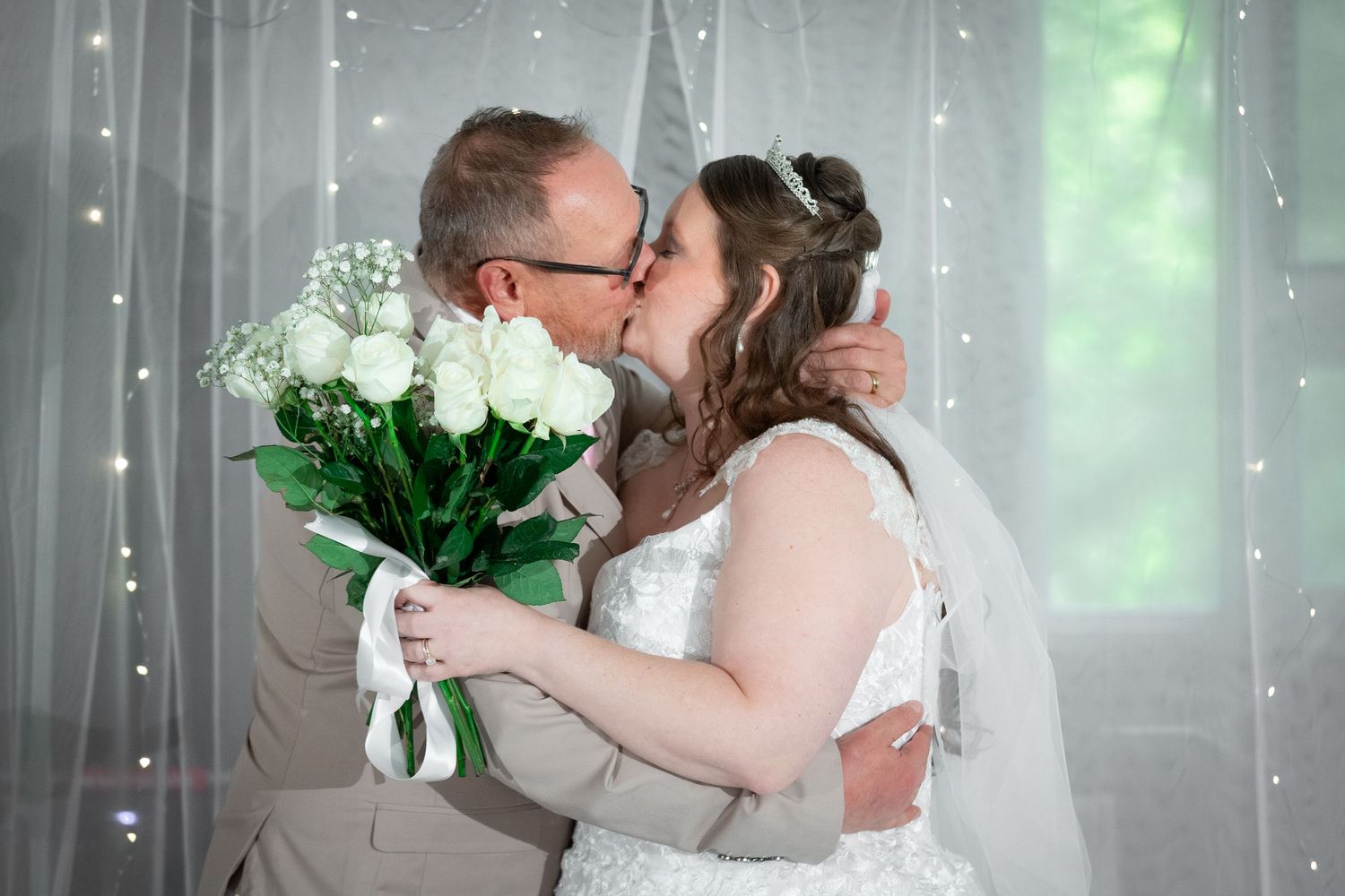 Newlyweds sharing a kiss while groom holds white rose bouquet at wedding ceremony.