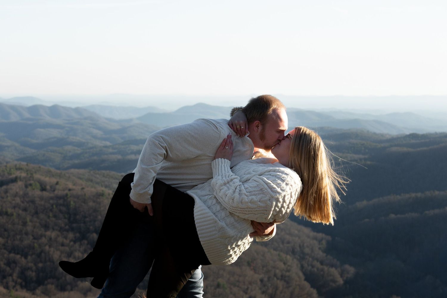 Couple shares romantic kiss on mountain overlook with scenic valley and rolling hills in background at sunset.