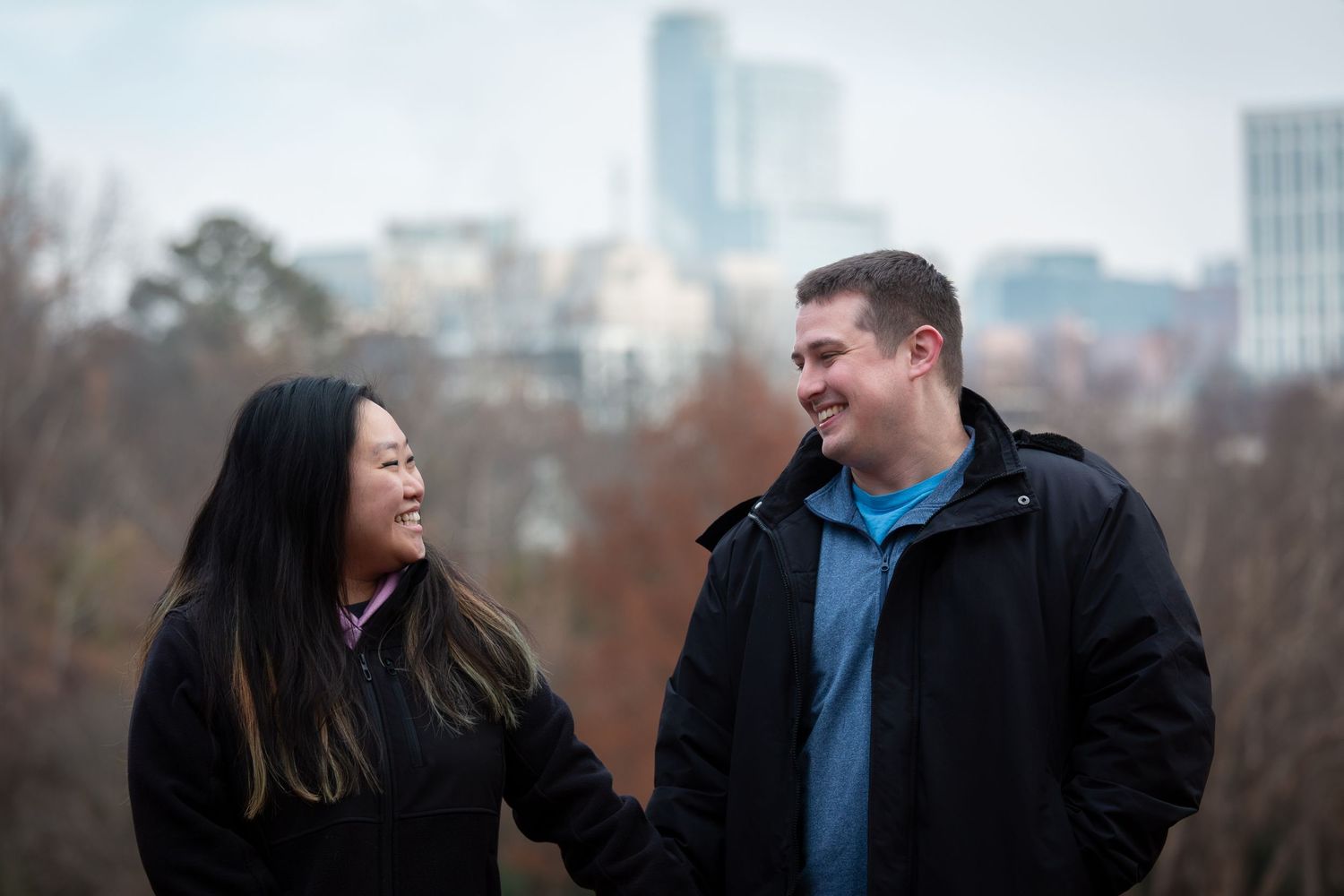 A couple shares a joyful moment together while walking outdoors on an autumn day with city buildings in the background.