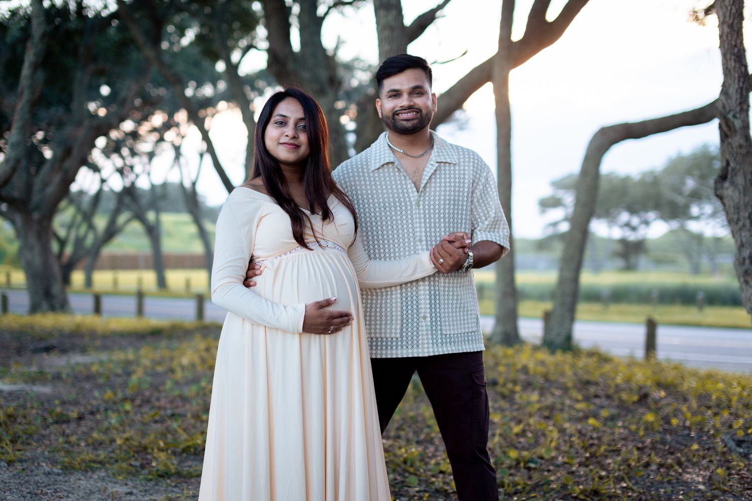A couple poses together outdoors in a romantic setting with trees and yellow flowers in the background.