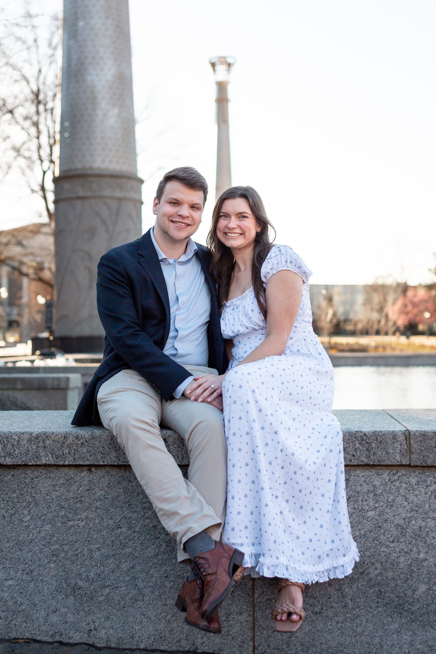 A couple sits together on a stone ledge near a decorative column during a sunny outdoor photo session.