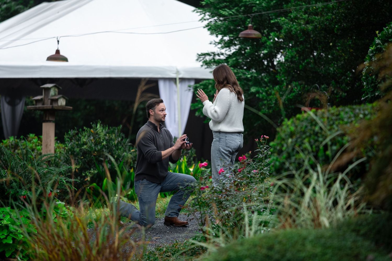 Man kneeling on one knee in a lush garden proposing to a woman standing near a white event tent.