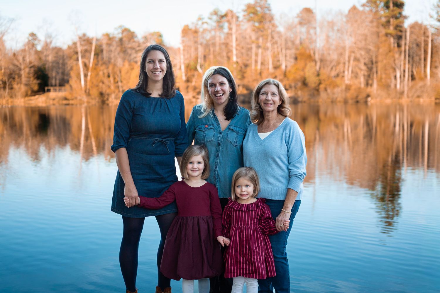 Family portrait of five women and girls smiling by a serene lake with autumn trees in the background.