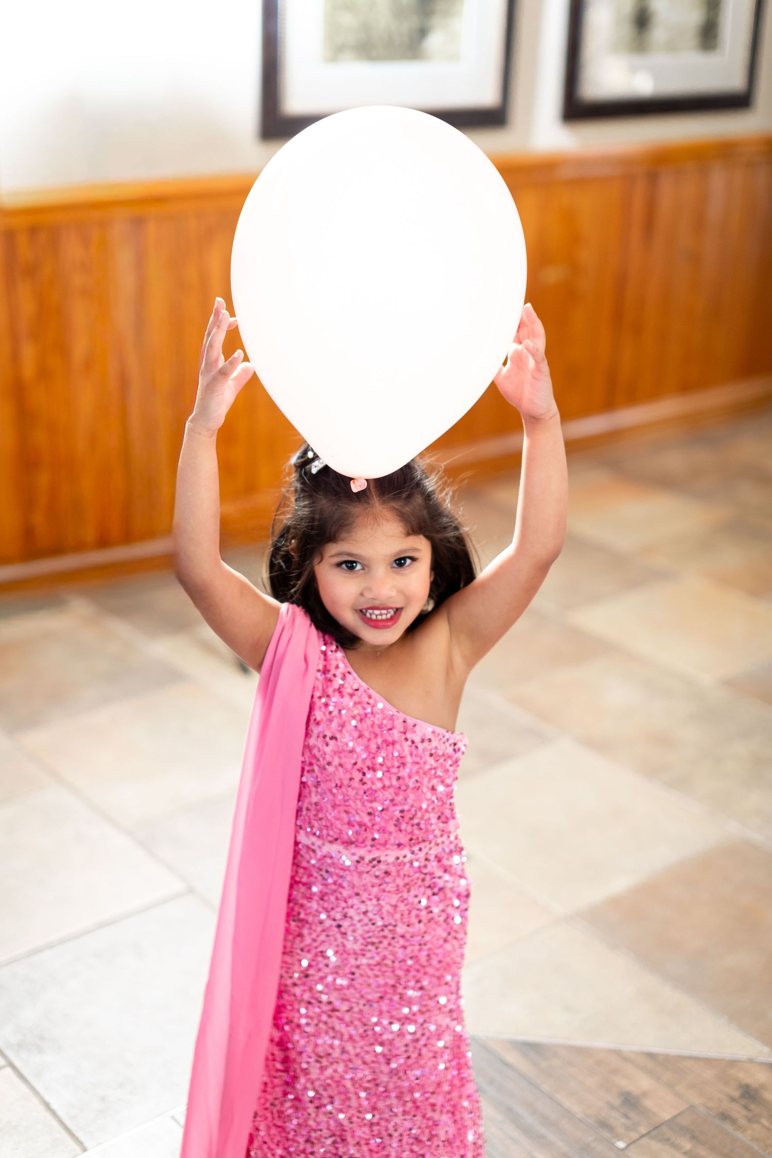 Child in a pink sequined dress plays with a white balloon in an indoor setting.