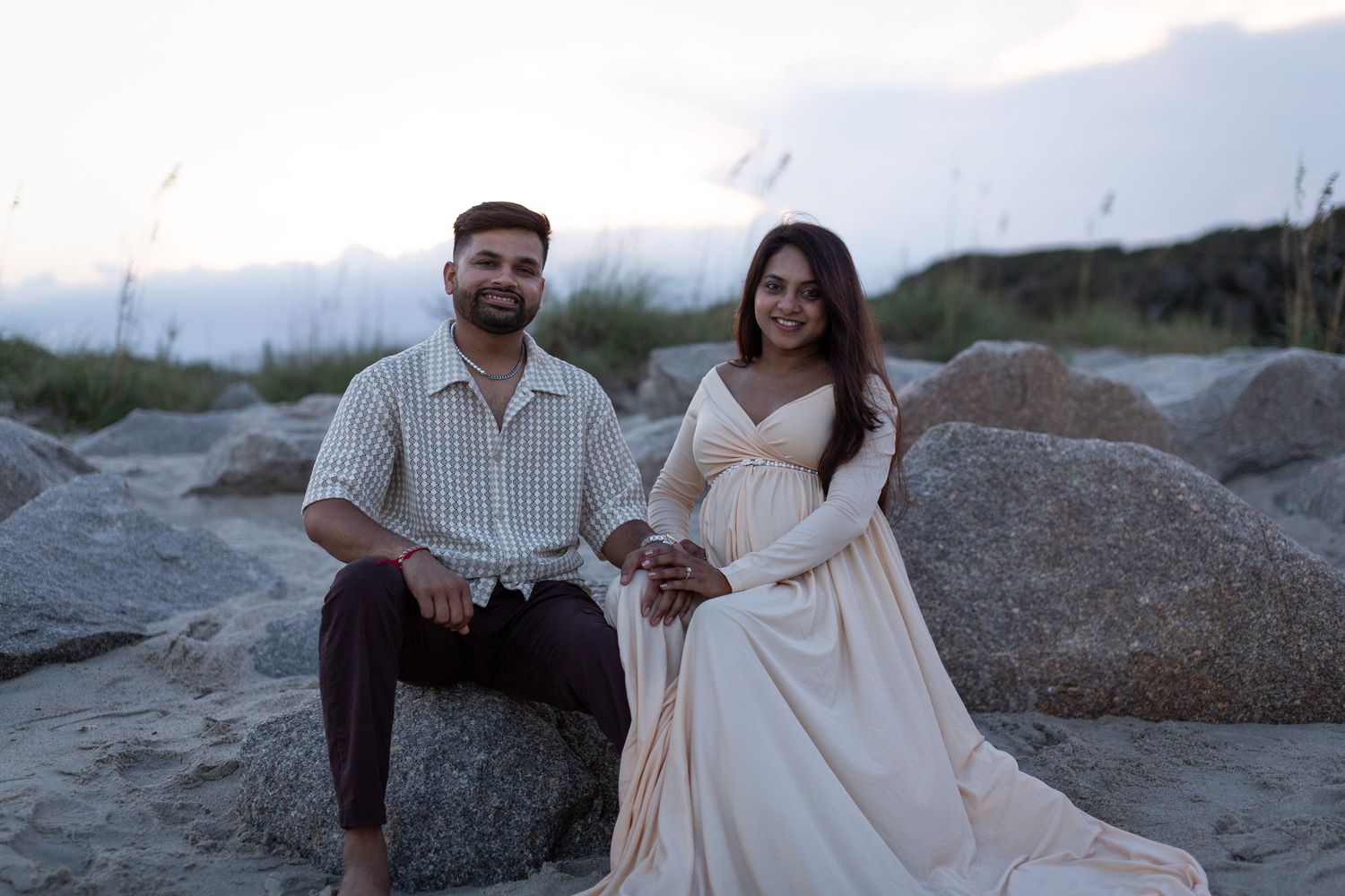 A couple in light colored outfits poses together on a rocky beach during sunset.