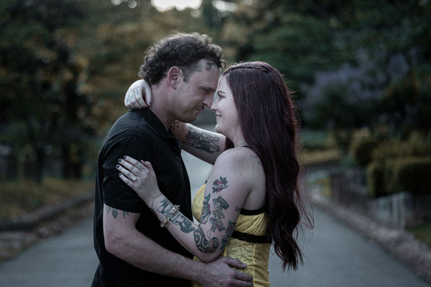A tattooed couple embraces intimately on a tree-lined path, gazing into each other's eyes in a romantic outdoor setting.