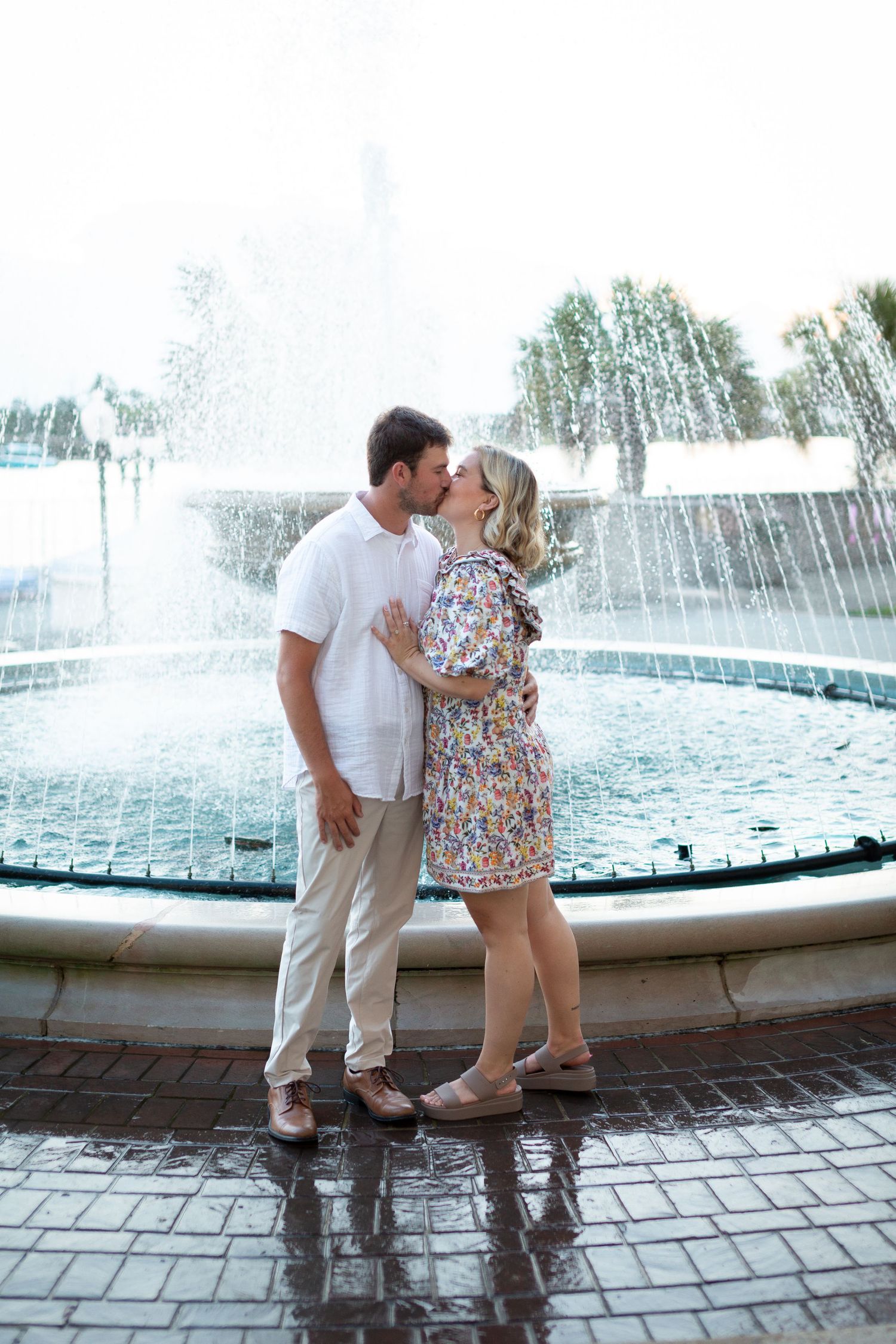 A couple shares a romantic moment by a fountain with water spraying in the background on a sunny day.