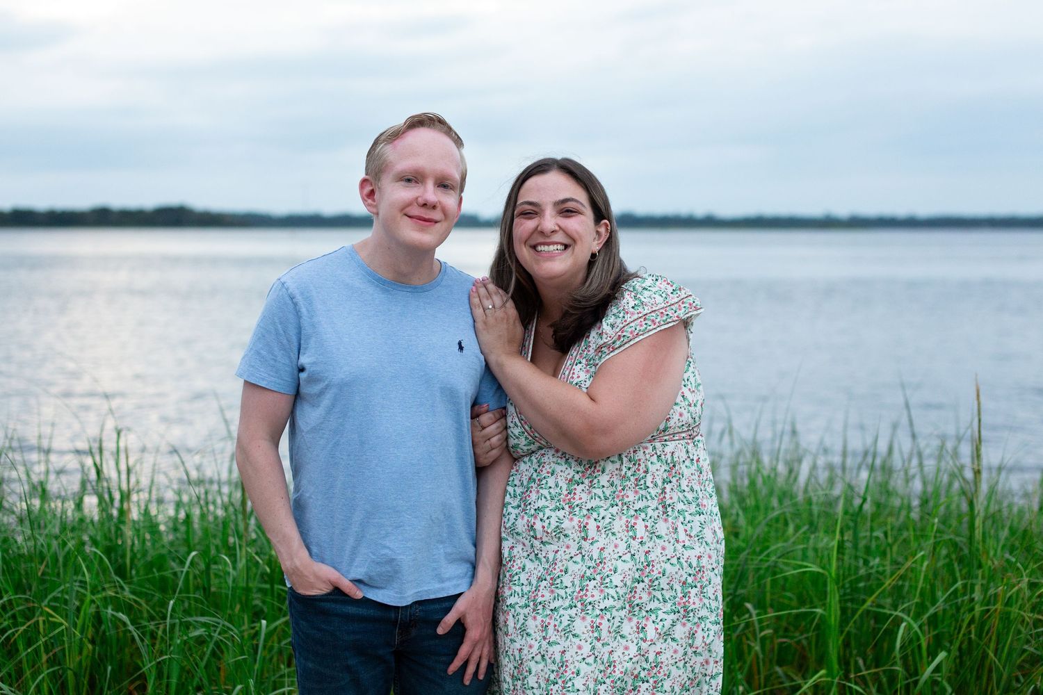 A couple stands together smiling by a lake at dusk with tall grass in the foreground.