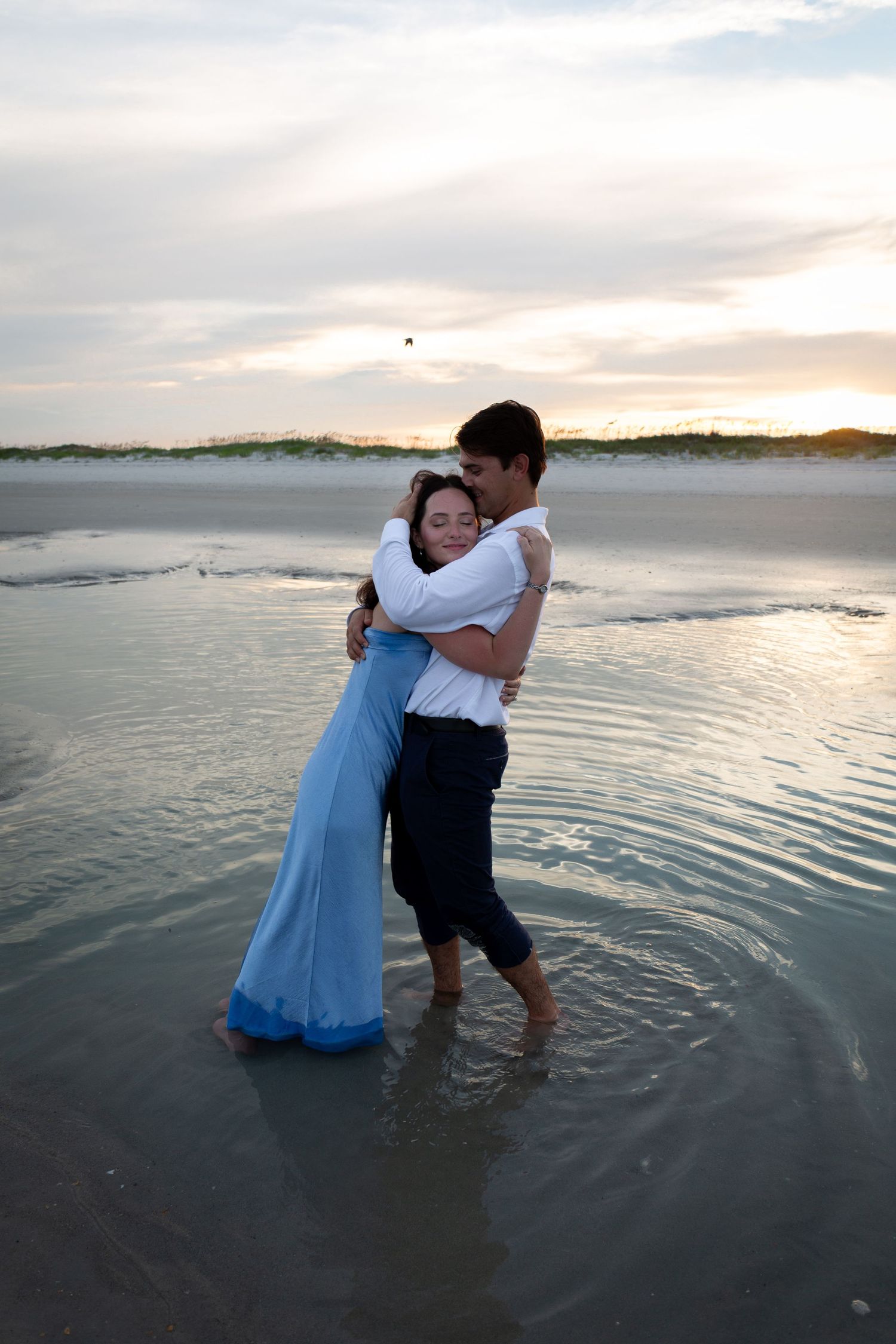 Couple embracing romantically in shallow ocean water at sunset, woman in blue dress, man in white shirt.