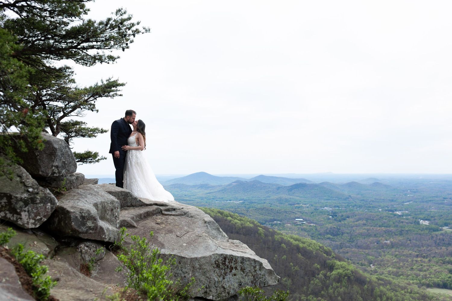 Bride and groom kissing on rocky mountain summit with sweeping valley views on their wedding day.
