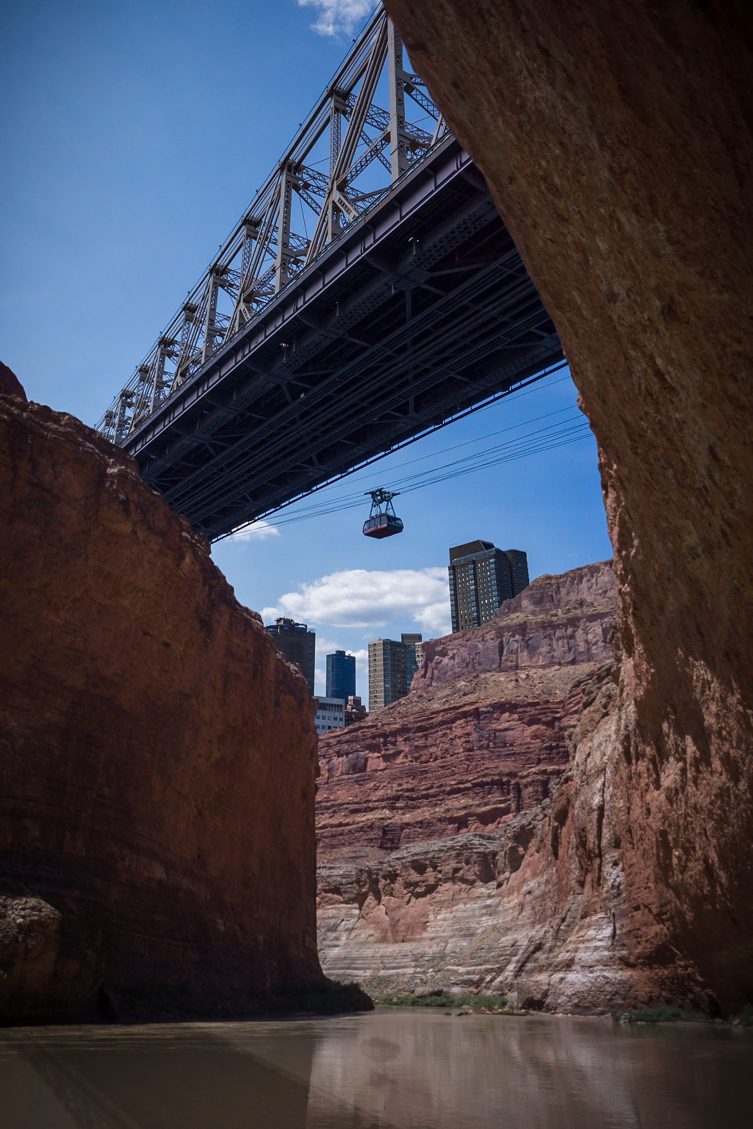 View from under a steel bridge in a red rock canyon with a cable car and city skyline.