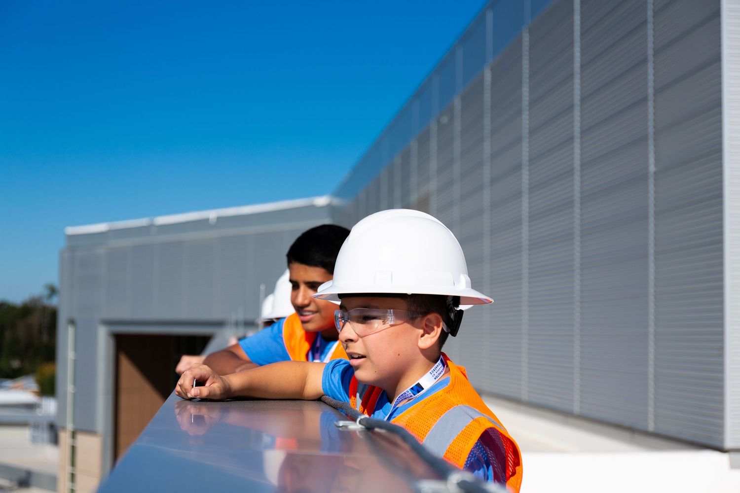 Construction workers in orange safety vests and hard hats inspect a building exterior with metal siding panels.