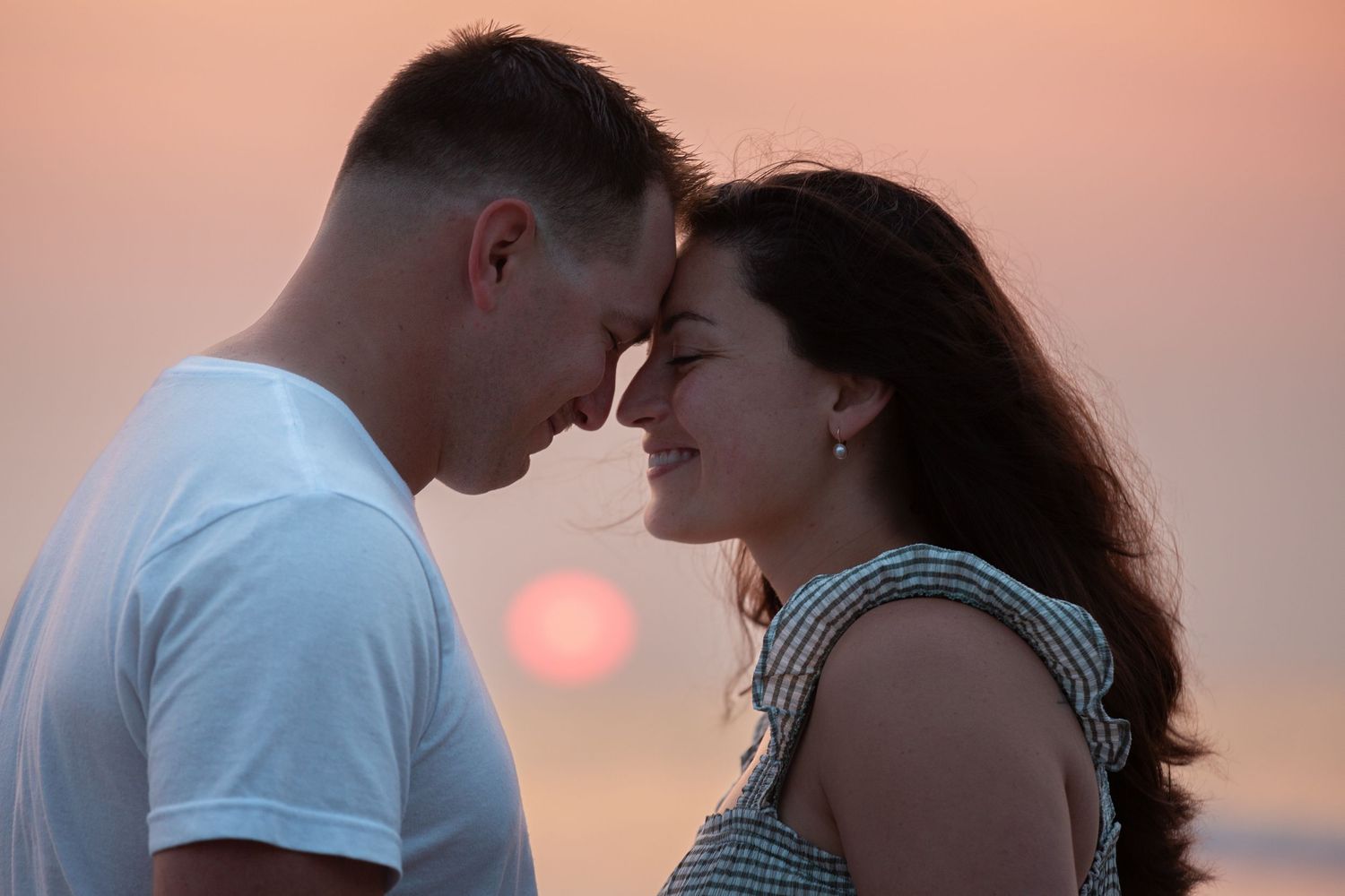 A romantic couple shares an intimate moment at sunset with an orange sun glowing in the background.