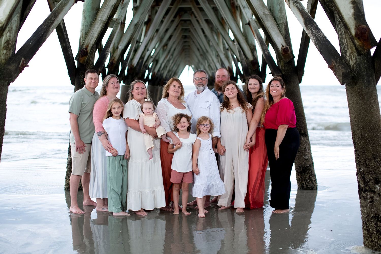 Extended family group photo under a wooden pier structure at the beach during sunset.