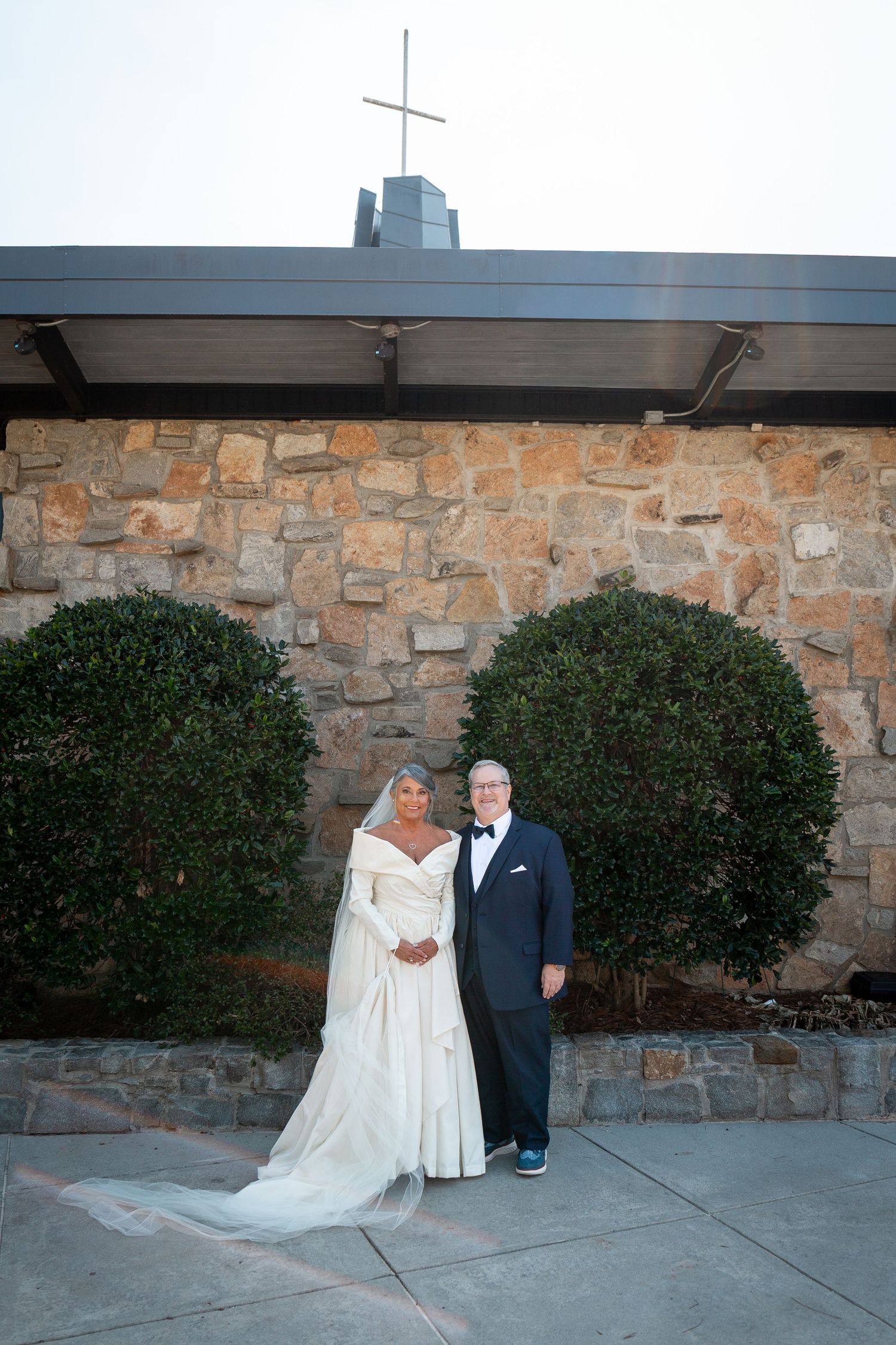 A wedding couple poses for portraits against a stone wall backdrop with decorative shrubs.