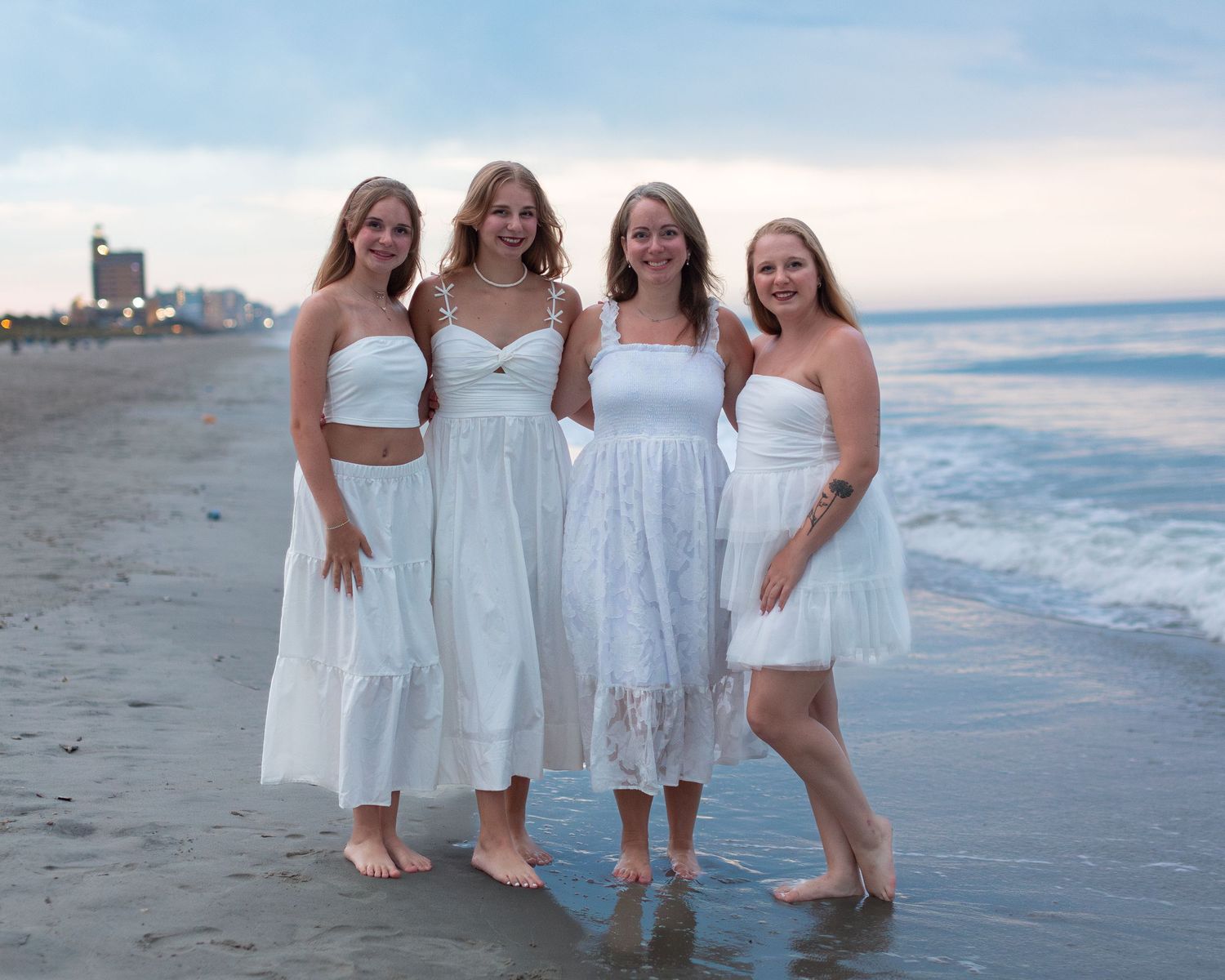 Four women in white dresses standing together on a beach at sunset.