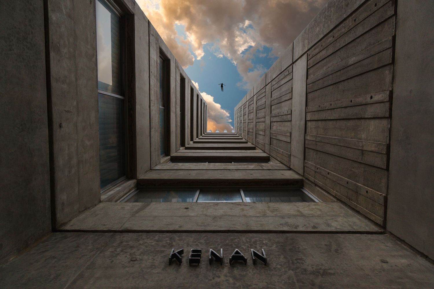 Low angle view looking up between concrete walls toward dramatic cloudy sky with Kenan text visible.