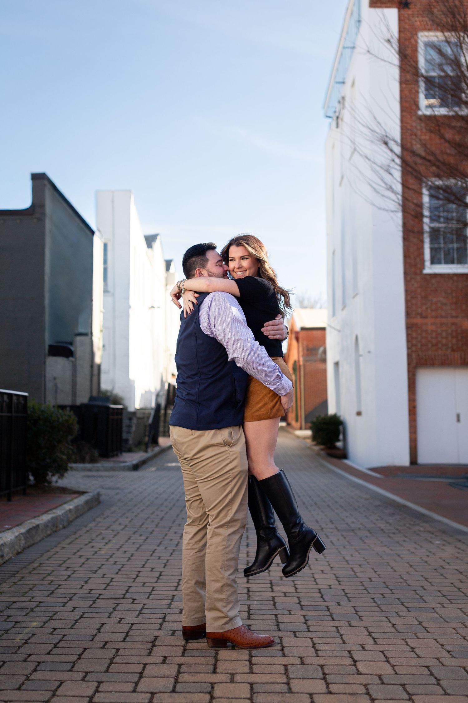 A romantic couple embraces and kisses on a brick paved walkway in front of white buildings.