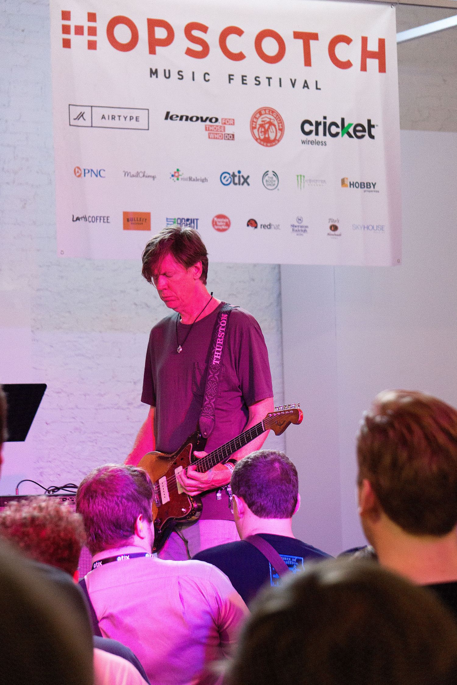 Musicians perform on stage at the Hopscotch Music Festival with pink lighting and sponsor banners visible in background.