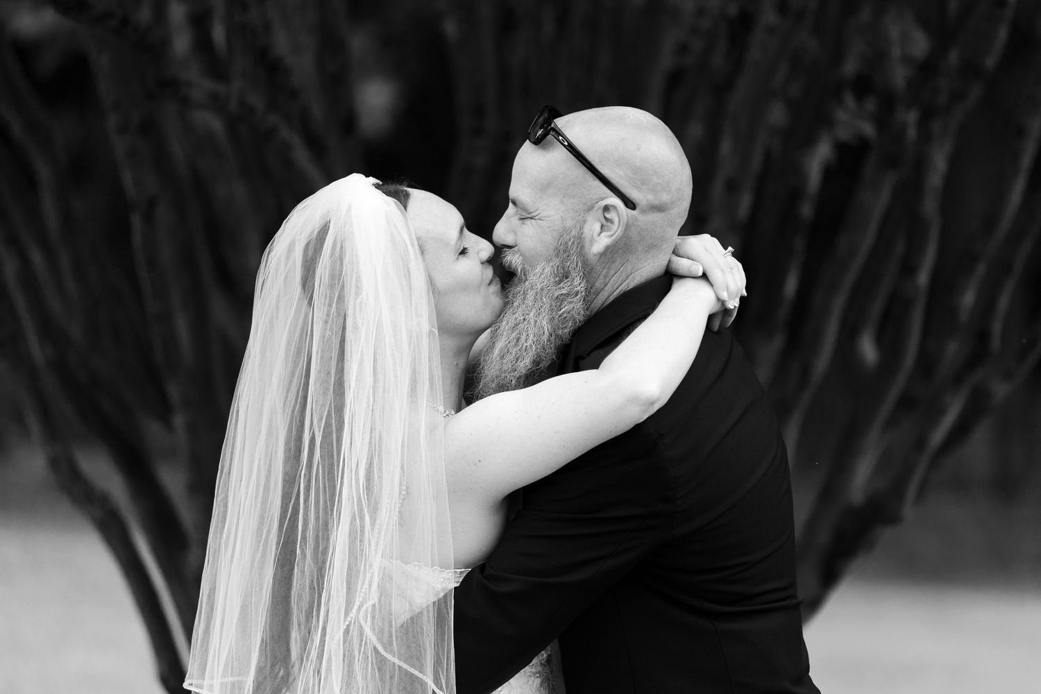 Black and white photo of a bride in a long veil embracing and kissing the forehead of a bearded groom with sunglasses on his head.