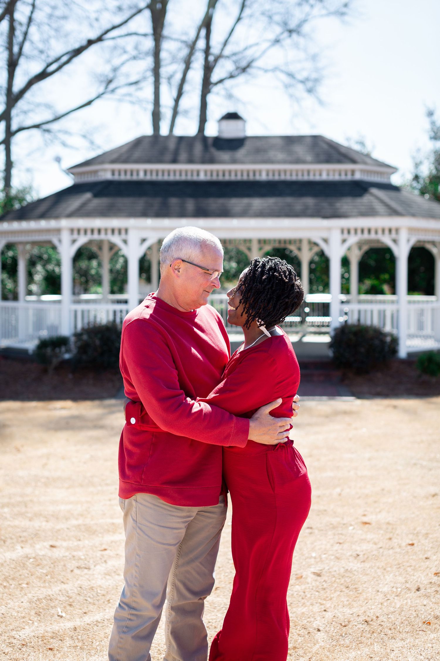A couple in matching red and khaki outfits embrace lovingly in front of a white gazebo on a sunny day.