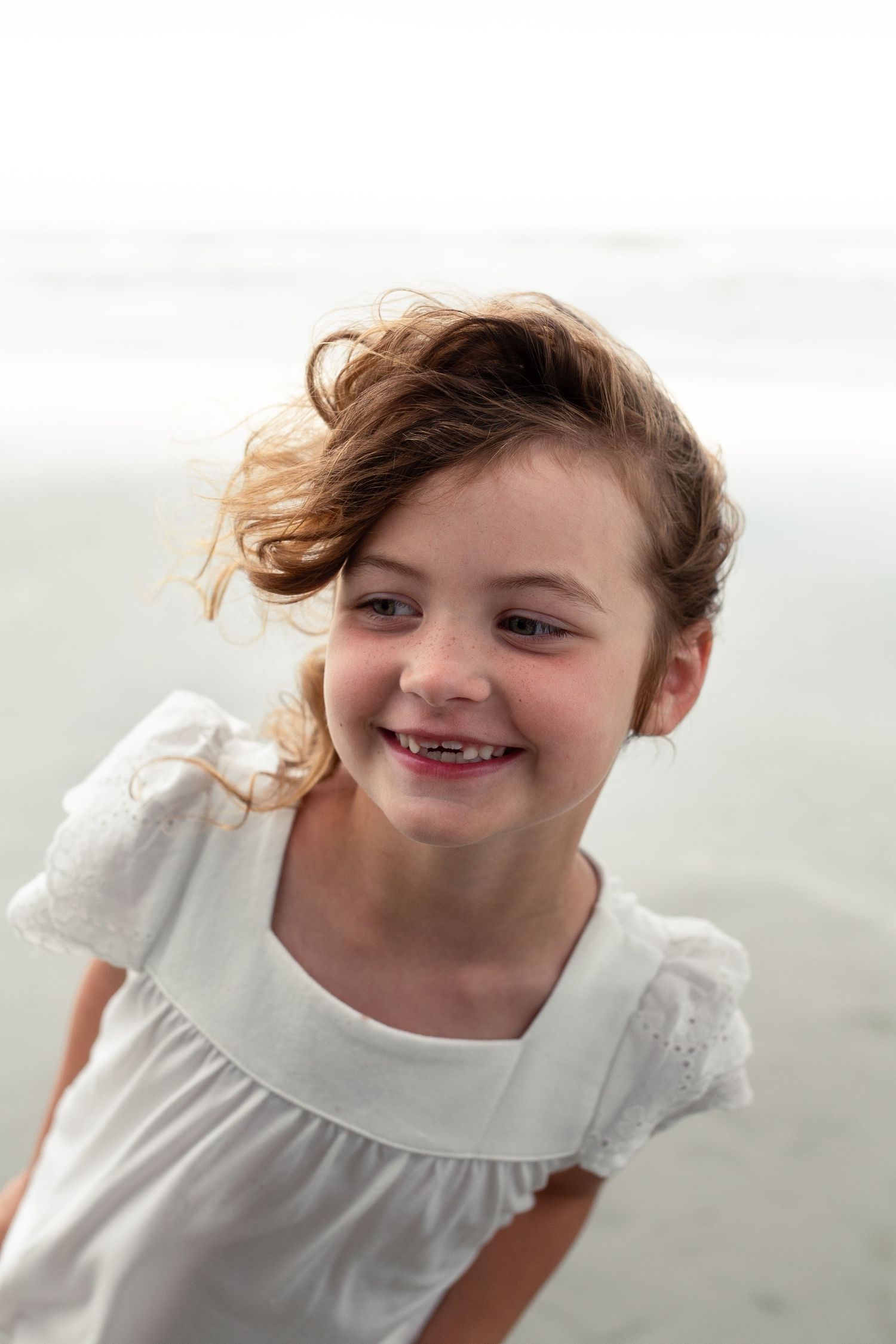 Young child in a white dress smiling with windswept hair against a light background.
