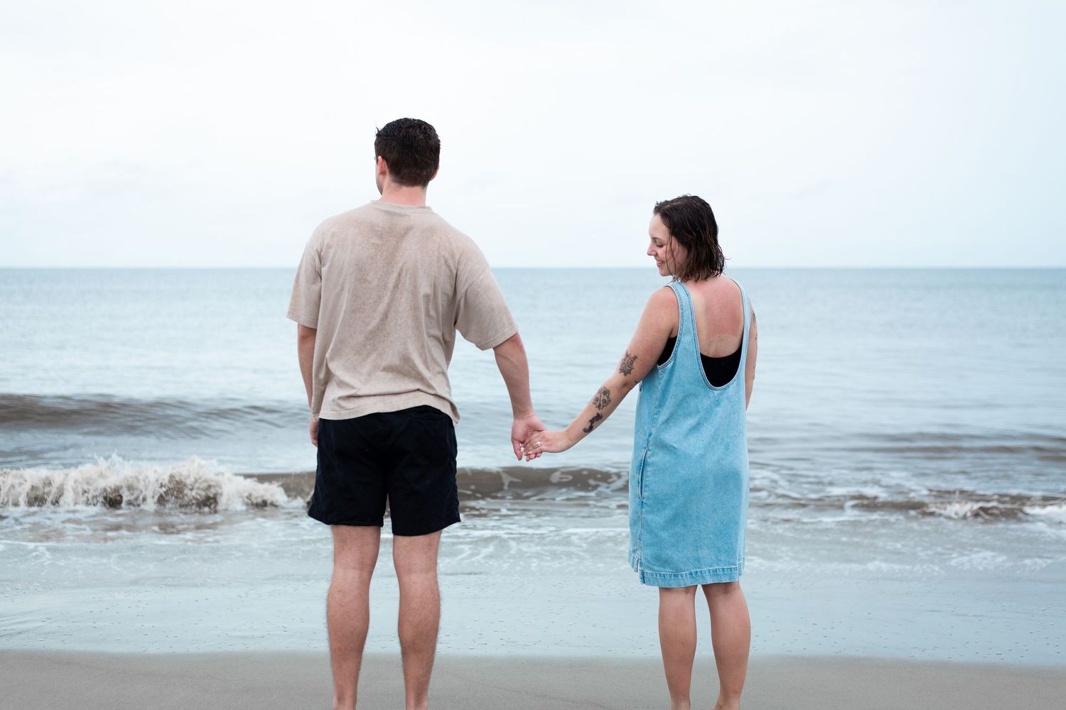 Two people walking along a misty beach shoreline holding hands on an overcast day.