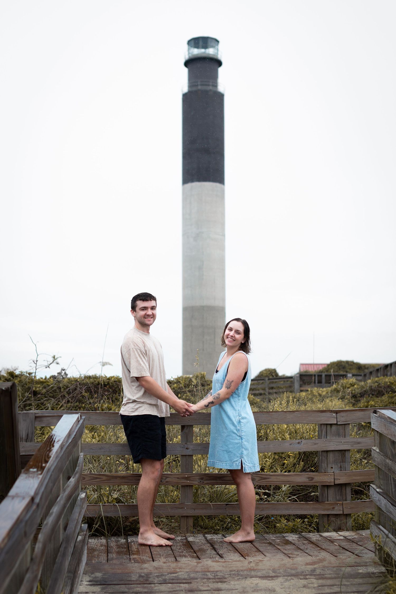 Two people stand on a wooden boardwalk near a lighthouse during what appears to be a marriage proposal.