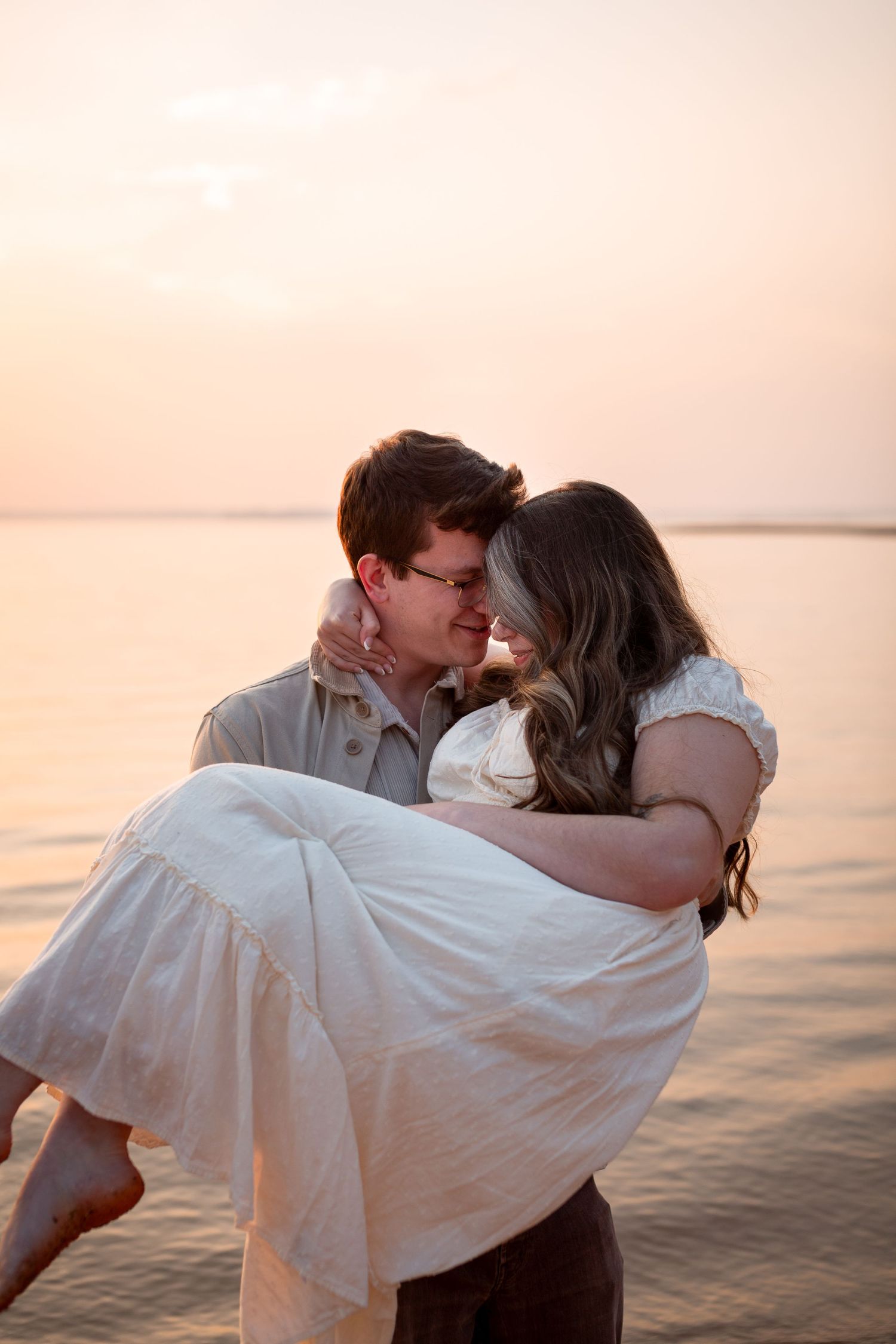 Romantic couple sharing a tender moment at sunset on the beach, man carrying woman in white dress.
