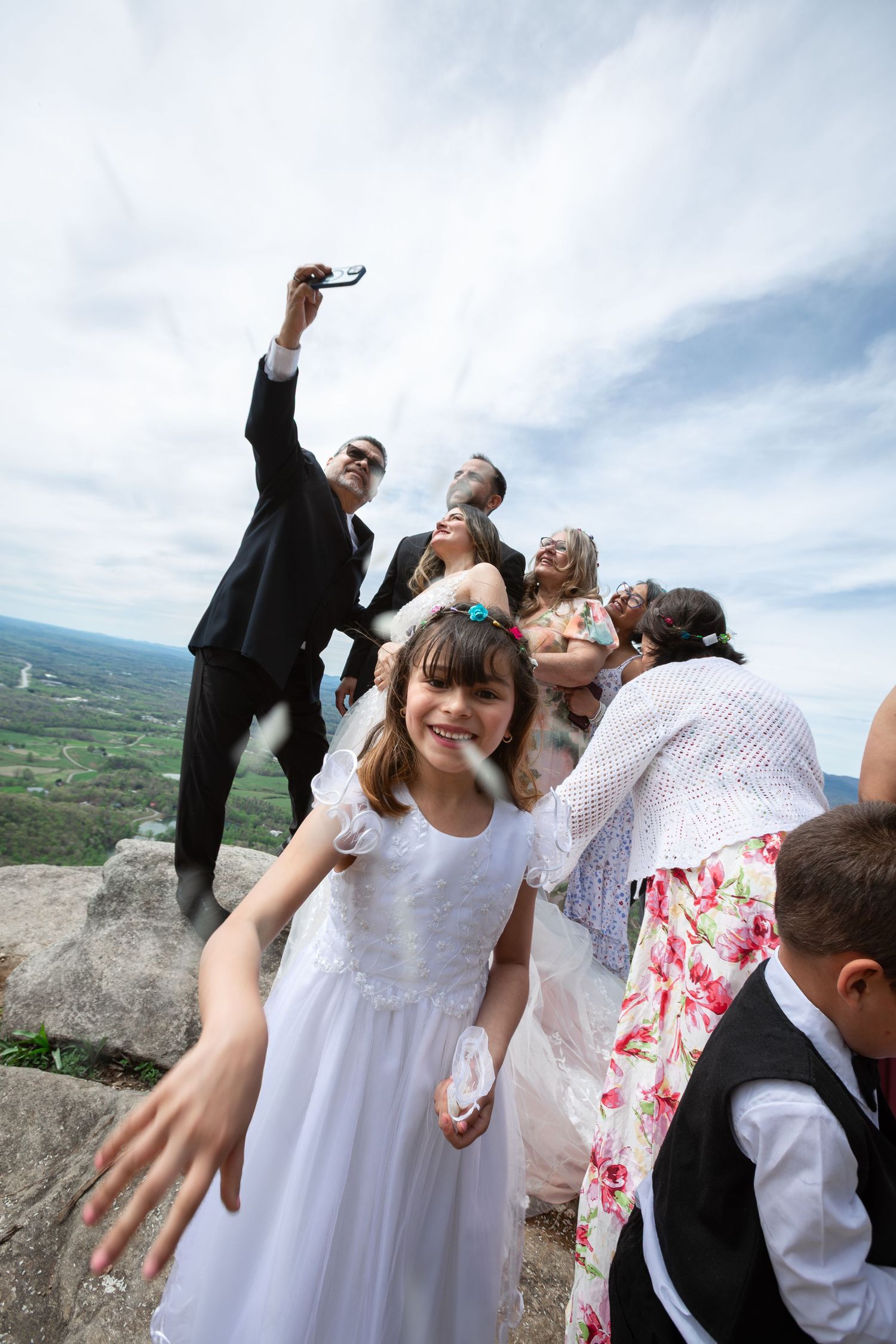 Girl in white dress smiles at camera during outdoor wedding celebration on rocky hilltop with guests in background.