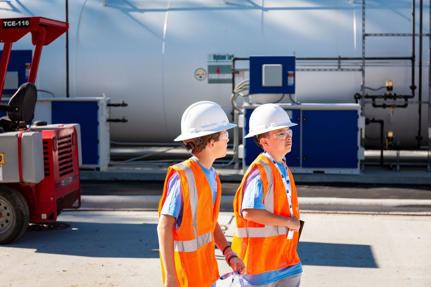 Industrial workers in safety gear inspect equipment at an outdoor facility with storage tanks and piping systems.
