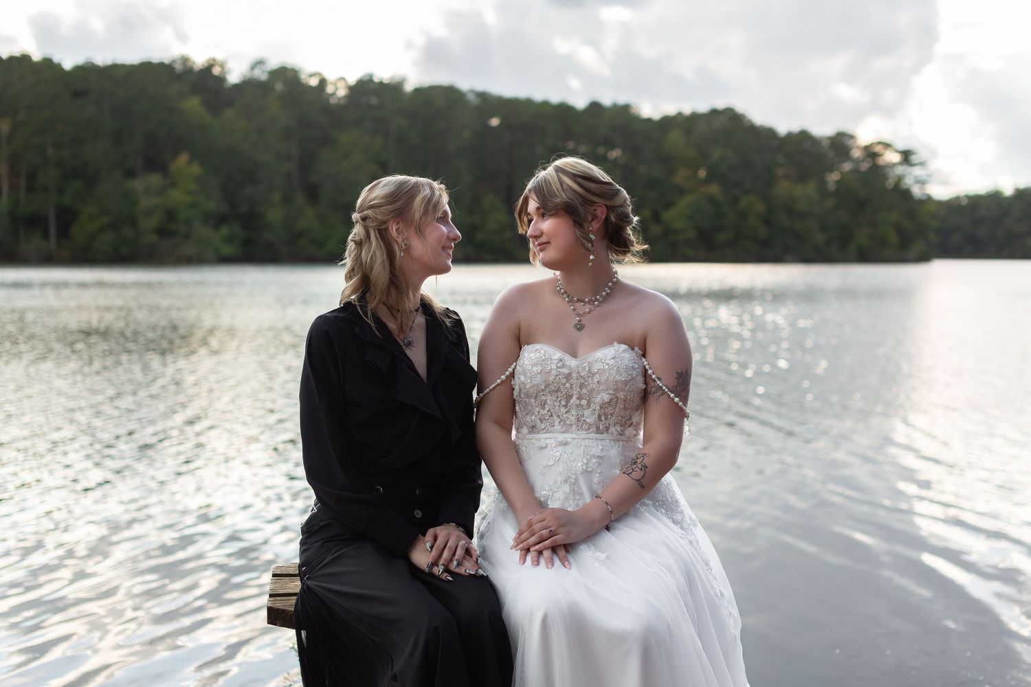 Two brides in a white lace gown and a black dress smile at each other while sitting on a dock beside a calm lake.