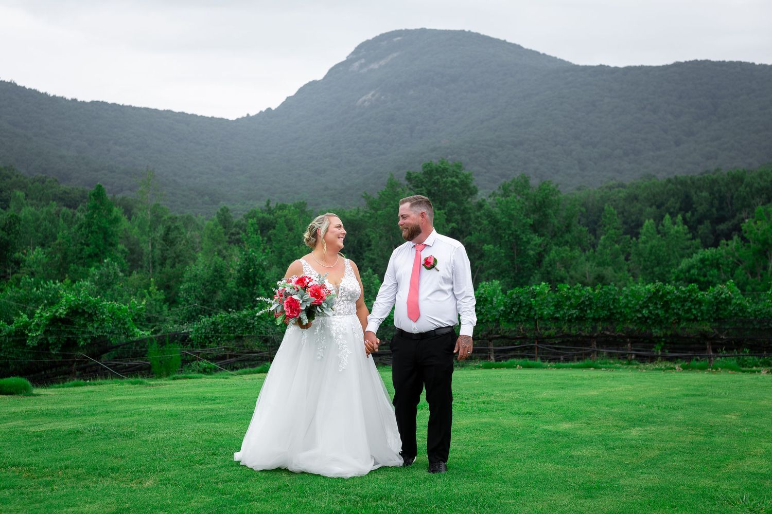 Bride in a white wedding gown holding a bouquet and groom in a pink tie stand hand in hand on a green lawn before forested mountains.