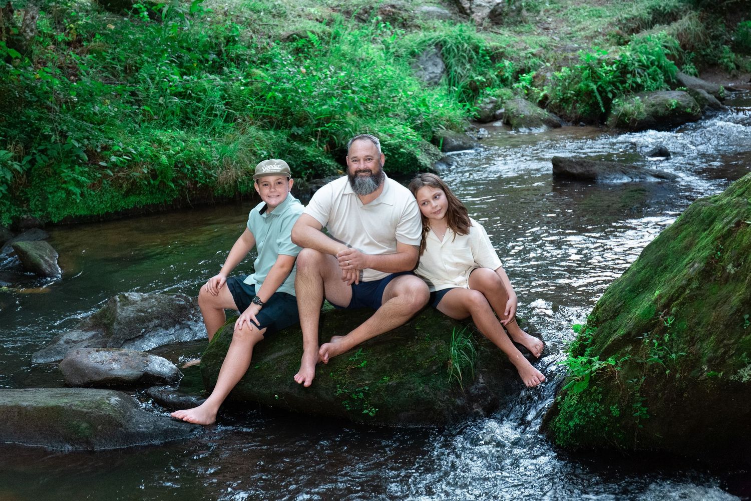 Father with a beard sits on a mossy rock with his son and daughter beside a peaceful forest stream.