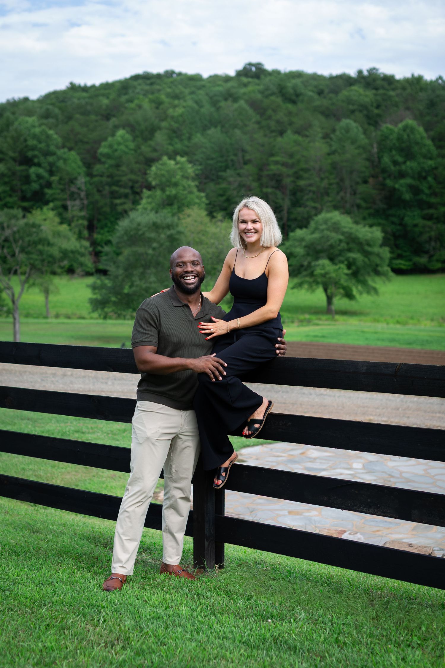 Happy interracial couple posing by a black wooden fence with lush green hills in the background.