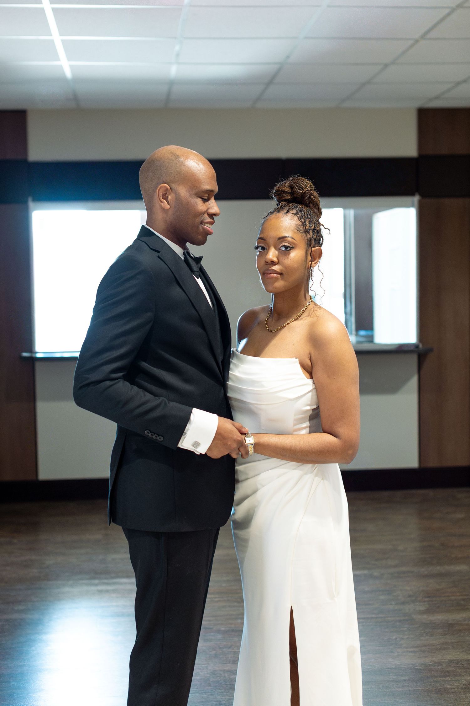 A couple in formal attire dances together in an empty indoor space with large windows.