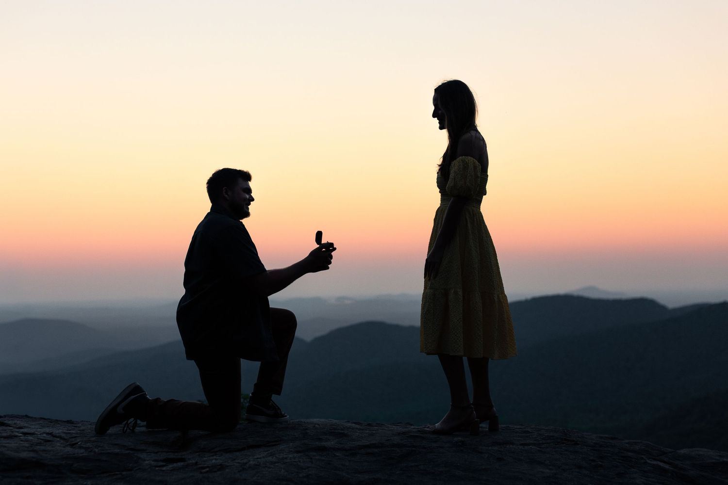 Silhouette of man on one knee proposing to woman at sunset on mountain peak with colorful sky backdrop.