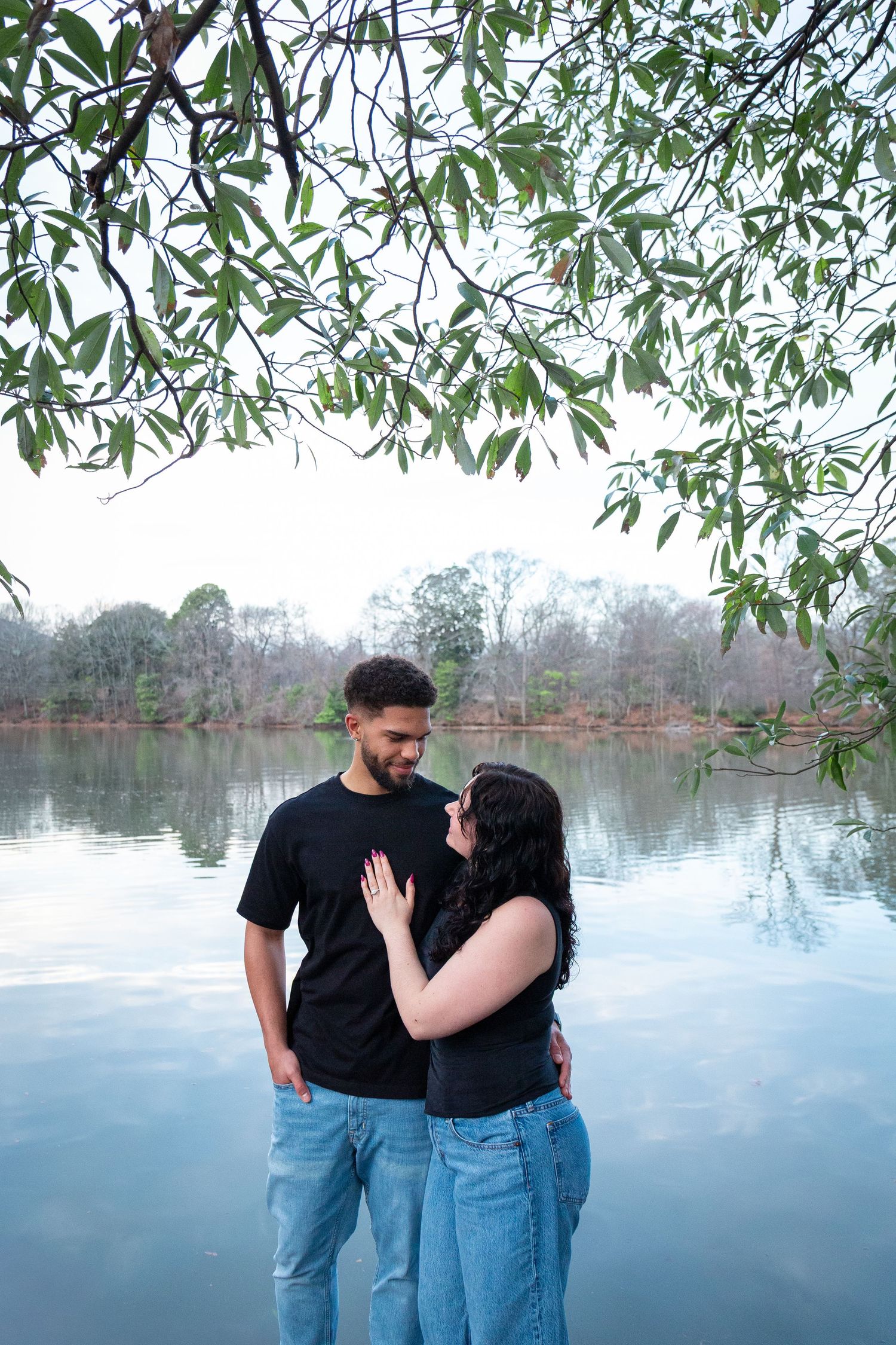 A couple shares an intimate moment by a serene lake with overhanging willow branches framing the scene.