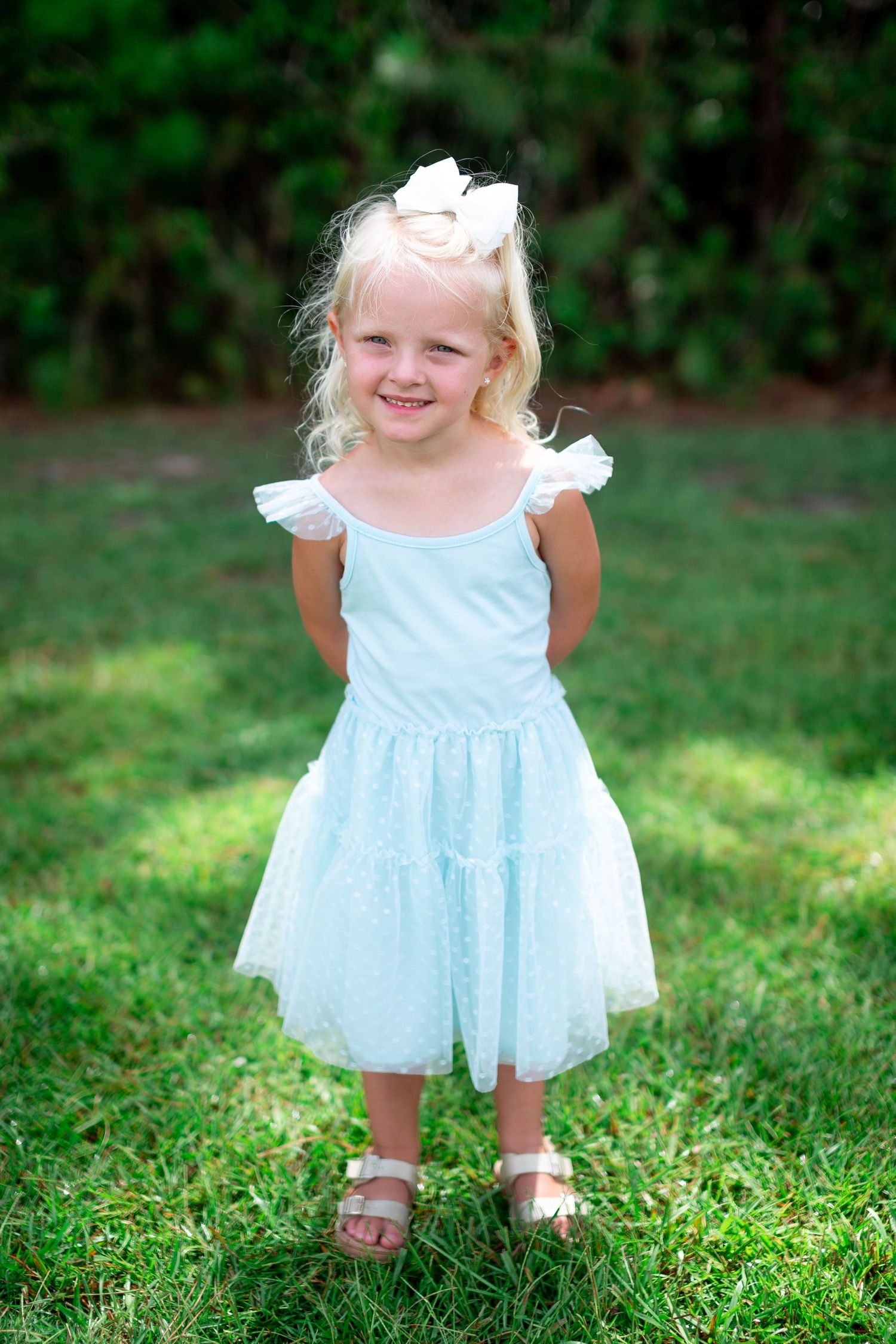 A young child in a light blue tulle dress stands smiling on grass on a sunny day.