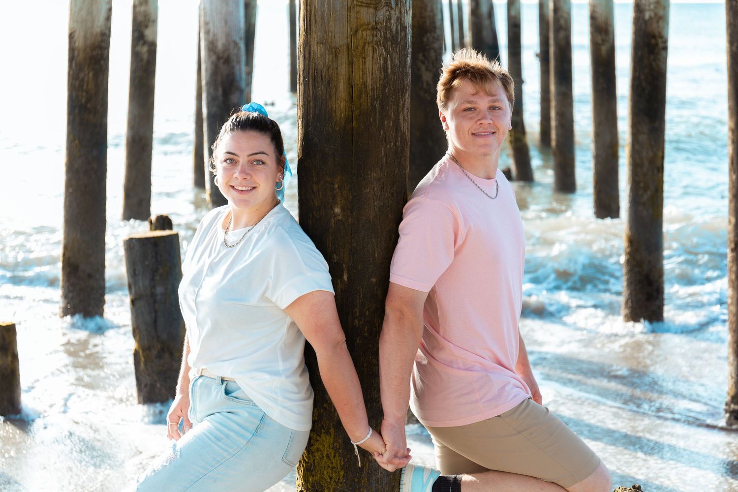 Young couple holds hands while leaning against a wooden pier piling with ocean waves washing around them.