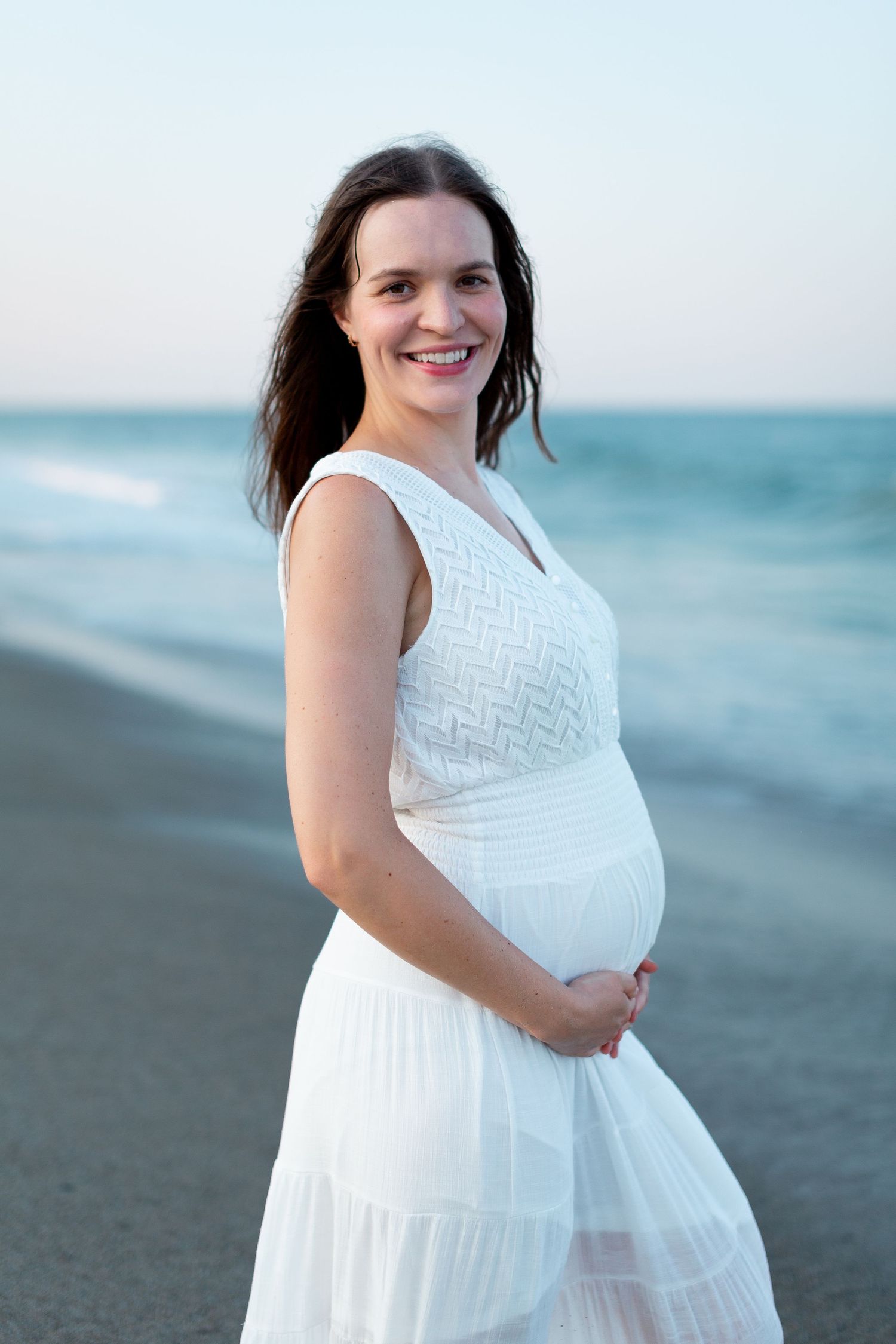 A person in a white dress poses for maternity photos on a beach with soft blue ocean waves in the background.