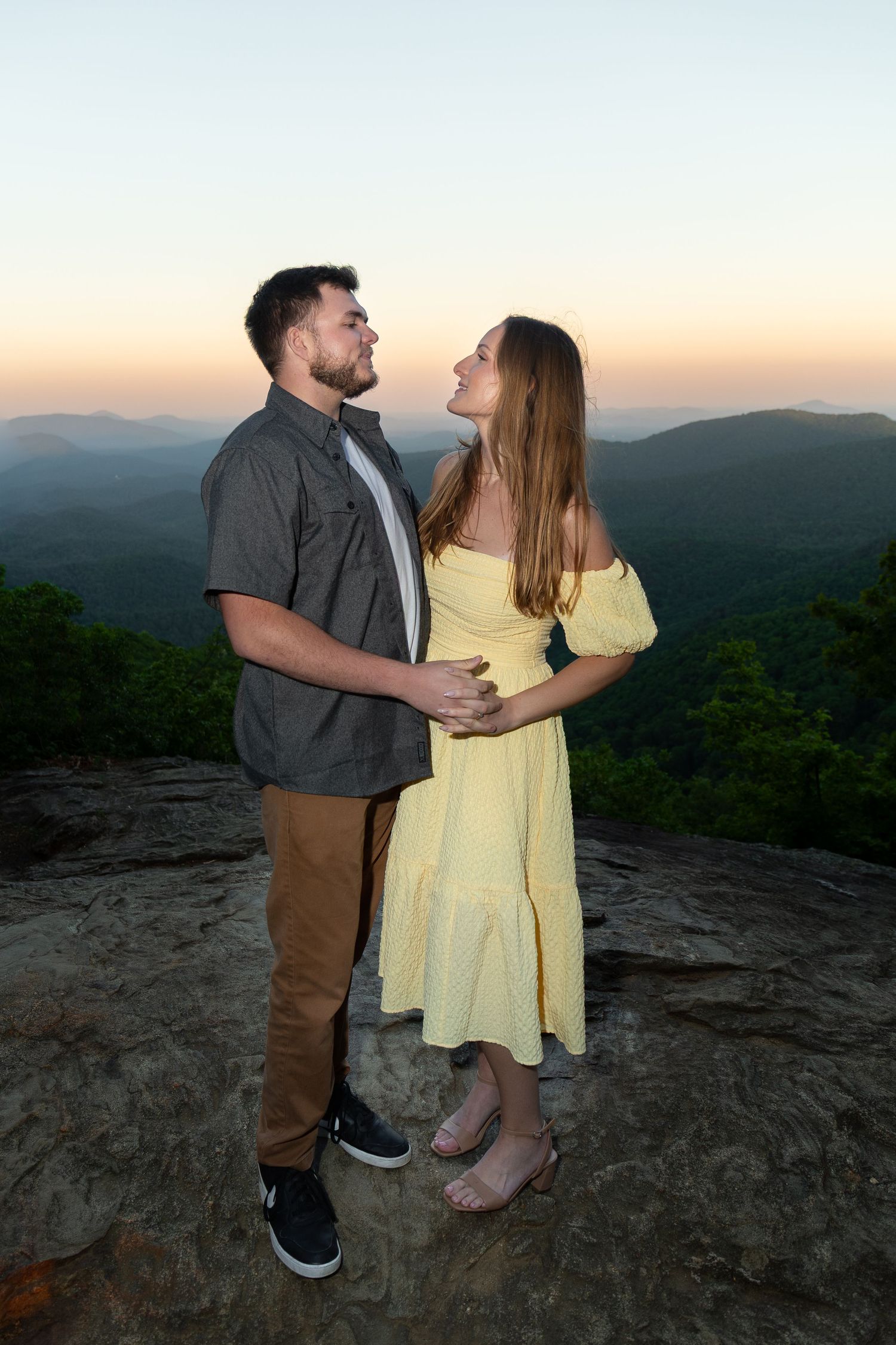 Couple embracing on rocky mountain overlook at sunset, woman in yellow dress gazing up at man in gray shirt.