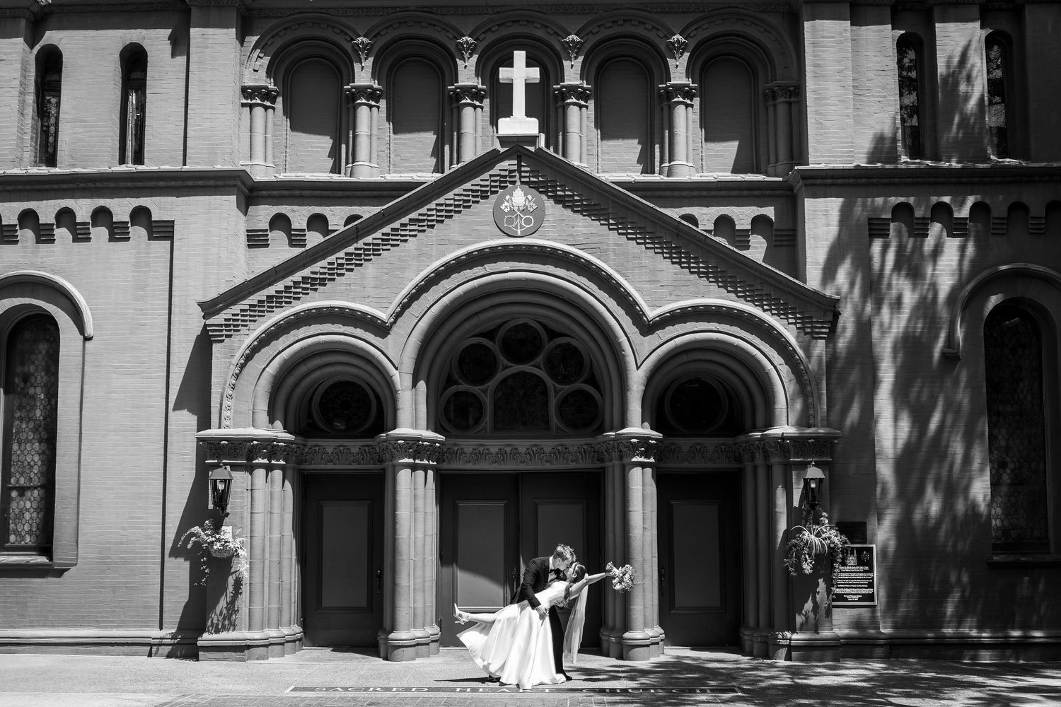 A black and white wedding photo of a bride and groom in a romantic dip in front of a grand church entrance with a cross.