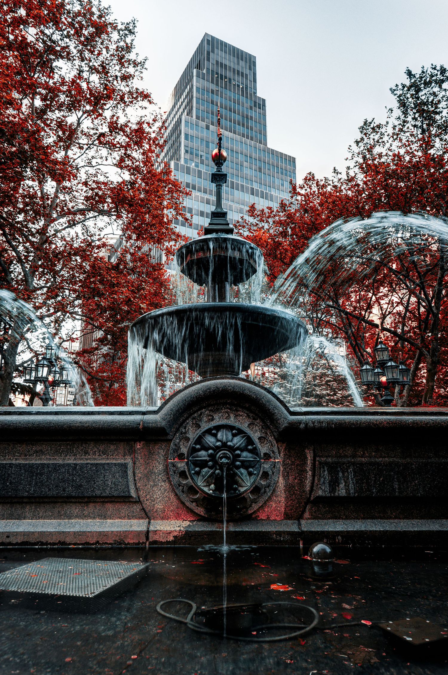 Ornate tiered fountain with red autumn trees and city skyscraper backdrop.